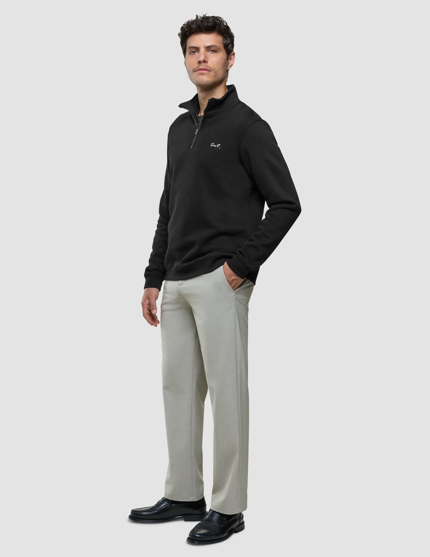 Ein Mann, der ein schwarzes Sweatshirt mit halbem Reißverschluss und der Stickerei „Smt.“ auf der Brust, eine hellgraue Hose und schwarze Slipper trägt.